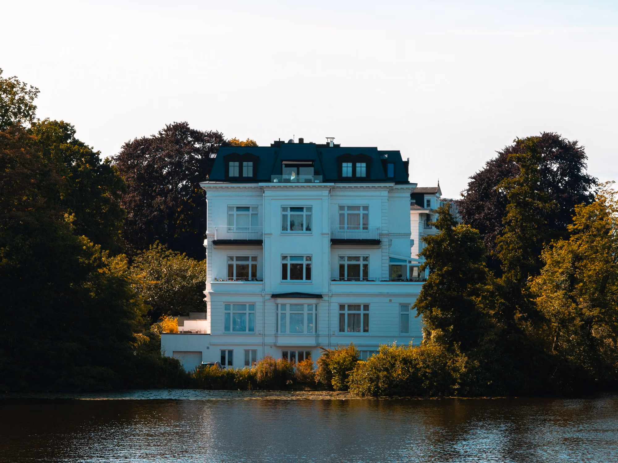 Weiße Stadtvilla mit Mansarddach an einem Gewässer, umgeben von Bäumen